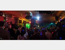 Eine belebte Bar oder Clubszene mit zahlreichen Menschen, die unter einer glitzernden Discokugel tanzen und feiern. Der Raum ist mit bunten Lichtern beleuchtet, und an der Decke hängen weihnachtliche Dekorationen.