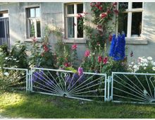 Ein steinernes Haus mit einfachen weißen Fenstern, vor dem ein üppiger Garten mit bunten Blumen, darunter blaue Rittersporne und rote Rosen, angelegt ist. Ein dekorativer, weißer Metallzaun grenzt den Garten ab, im Vordergrund ist eine Rasenfläche sichtbar.