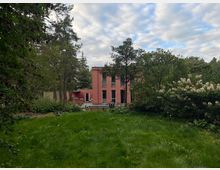 Moderne zweigeschossige Villa in rosafarbener Fassade, umgeben von einer grünen Wiese, Bäumen und Büschen. Der Himmel ist teils bewölkt und das Haus befindet sich in einer ruhigen, naturnahen Umgebung.