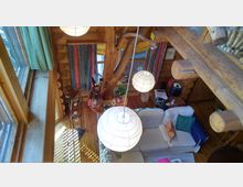 Holzinterior eines gemütlichen Wohnzimmers mit sichtbaren Holzbalken und -wänden, bunten Vorhängen, weißen Papierlampen und einer Sofaecke mit bunten Kissen. Ein Baumstamm steht in der Mitte des Raumes, und durch große Fenster fällt Tageslicht ein.