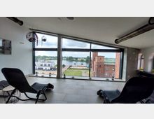 Innenraum eines modernen Gebäudes mit großen Fenstern, die einen Blick auf eine Flusslandschaft, ein Ziegelgebäude und moderne weiß gestrichene Häuser bieten. Im Vordergrund stehen Liegestühle und kleine Dekorationen auf der Fensterbank.