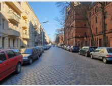 Eine gepflasterte Straße in einer Stadt mit parkenden Autos auf beiden Seiten. Links stehen beige Wohngebäude mit Balkonen, rechts ein großes Gebäude aus rotem Backstein mit hohen Fenstern und kahlen Bäumen entlang des Gehwegs.