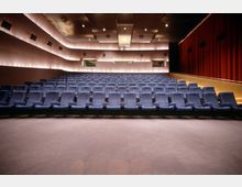 Kinosaal mit mehreren Reihen von gepolsterten, blauen Kinositzen in ansteigender Anordnung. Wände mit indirekter Beleuchtung, rote Vorhänge rechts und eine Decke mit eingebauten Leuchten dominieren die moderne Innenarchitektur.