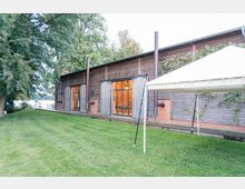 Holzgebäude mit großen Glasfenstern und Schiebetüren, umgeben von einer gepflegten Rasenfläche und Bäumen; rechts steht ein offenes Zelt auf der Wiese, im Hintergrund ist ein Gewässer sichtbar.