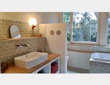 Modernes Badezimmer mit einem rechteckigen Waschbecken auf einer Holzablage, einer Ablagefläche mit beigefarbenen Fliesen und einem Wandspiegel. Im Hintergrund große Fenster mit Blick auf Bäume und ein Bereich mit einer Badewanne, ebenfalls gefliest.