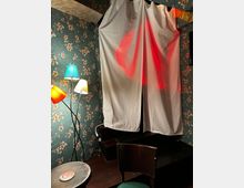 Ein gemütlicher Innenraum mit gemusterter Tapete in Blumenoptik, einem Vorhang, der rote Lichtreflexe zeigt, und einer Stehlampe mit bunten Lampenschirmen. Im Vordergrund befinden sich ein kleiner Tisch und ein Stuhl.