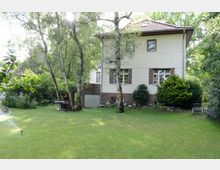 Zweistöckiges Wohnhaus mit heller Fassade und braunen Backsteinelementen, umgeben von einem großen Garten mit gepflegtem Rasen, Bäumen und Sträuchern. Im Garten ist ein Sprinkler aktiv, und es stehen Gartenmöbel unter den Bäumen.