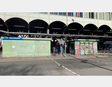 Ansicht des Görlitzer Bahnhofs in Berlin, mit einem erhöhten Eisenbahnviadukt und einer darüber verlaufenden Bahnstrecke. Im Vordergrund stehen ein grün gekacheltes Gebäude und ein kleiner Kiosk mit bunten Werbeplakaten sowie abgestellten Fahrrädern.