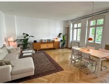 Helles Wohn- und Esszimmer mit Fischgrätparkett, großer beigefarbener Eckcouch auf gemustertem Teppich und Holzboden. An der Wand steht ein niedriges Sideboard mit Büchern und Lampe, daneben Zimmerpflanzen; rechts ein Esstisch mit Stühlen vor hohen Fenstern mit Blick ins Grüne.