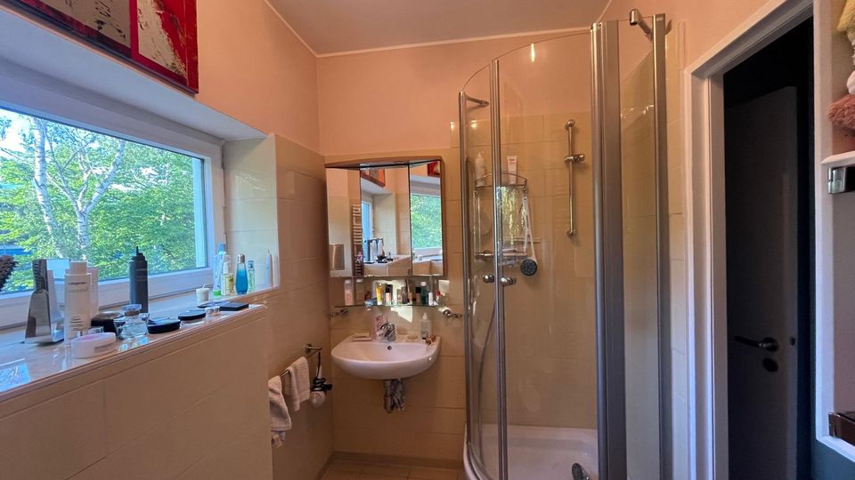 Kleines Badezimmer mit beige gefliesten Wänden, einem Waschbecken mit Spiegel und einer halbrunden Eckdusche mit Glasabtrennung. Links sorgt ein großes Fenster über einer Ablage mit Pflegeprodukten für Tageslicht und Blick ins Grüne.
