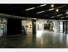 Eingangsbereich eines öffentlichen Gebäudes mit einem offenen und hellen Marmorboden, Treppen zu einem oberen Bereich rechts und Glastüren links. An den Wänden hängen Bilder, und Beleuchtung sowie Schilder wie „Garderobe“ und „Planetarium“ sind sichtbar.