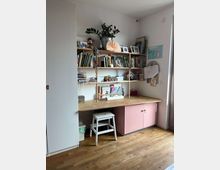 Arbeitszimmer mit einem Holztisch und einer weißen Sitzbank, darüber ein Regal mit Büchern, Pflanzen, Bildern und Bastelmaterial. Der Raum hat Holzboden, weiße Wände und eine Schrankecke links.