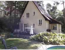 Zweistöckiges Haus mit heller Fassade und Satteldach, umgeben von Bäumen und einem Garten. Im Vordergrund eine Terrasse mit Gartenmöbeln, Sonnenschirm und einem kleinen Pool; umliegend Grünflächen und Farnpflanzen.