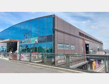 Bahnhof Warschauer Straße in Berlin mit moderner Fassade aus braunen Paneelen und großen Glasfronten. Vor dem Eingang befinden sich Werbetafeln, ein Geländer und darunter verlaufende Schienen sowie einige Personen auf der Brücke.