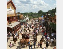 Ansicht einer belebten Westernstadt mit Holzgebäuden im Stil des Wilden Westens, zahlreichen Menschen auf der Straße, Pferden und Kutschen sowie Tipis im Hintergrund. Die Szenerie ist von Bäumen umgeben, und der Himmel ist leicht bewölkt.