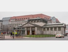 Historisches Gebäude mit Fassaden aus grauem Stein und grünem Dach, das als Eingangsbereich zu einer U-Bahn dient, daneben moderne Stadtarchitektur mit großen Gebäuden, darunter ein Kaufhaus mit rotem Dach. Im Vordergrund befinden sich Gehwege, ein Taxi und Personen, die die Straße überqueren.