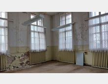 Ein leerer, verfallener Raum mit hohen Fenstern, durchsichtigen weißen Vorhängen und sichtbaren Heizkörpern unter den Fenstern. Die Wände sind stellenweise beschädigt, mit abblätternder Farbe und freigelegtem Mauerwerk.