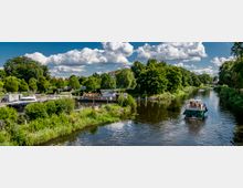 Flusslandschaft mit grünen Ufern, vereinzelt kleinen Booten und einem Hausboot am Rand. Im Hintergrund sind Bäume, Wiesen und einige Gebäude unter einem Himmel mit weißen Wolken zu sehen.
