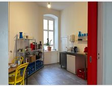 Kleine Küche mit weiß gestrichenen Wänden, einem großen Fenster, einem Metallregal mit Küchenutensilien und einem Tisch mit gelben Stühlen. Auf der rechten Seite befinden sich eine Spüle, ein Geschirrspüler und weitere Küchengeräte, umgeben von modernen Fliesen.