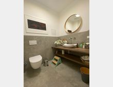 Modernes Badezimmer mit grauen Fliesen, hängender Toilette und Holzwaschtisch. Ein runder Spiegel hängt über dem Waschtisch, und eine kleine grüne Kindertrittleiter steht darunter.