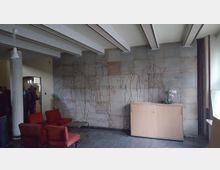 Innenraum mit einer Wandmalerei, die stilisierte Umrisse von historischen Persönlichkeiten zeigt, deren Namen und Lebensdaten angegeben sind, darunter Michael Faraday und James Clerk Maxwell. Im Vordergrund stehen rote Polsterstühle und ein Glastisch, daneben ein Holzschrank mit einer weißen Orchidee als Dekoration.