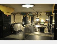 U-Bahn-Station mit moderner Architektur, Rolltreppen, glänzenden schwarzen Wandflächen und geometrisch gestalteten gelben und grünen Deckenleuchten. Im Hintergrund ist ein Schild mit der Aufschrift „nur Ankunft“ sichtbar.