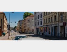 Straßenszene in einer urbanen Umgebung mit mehrstöckigen Wohn- und Geschäftsgebäuden auf beiden Seiten. Eine Straßenbahnlinie verläuft entlang der Straße, flankiert von parkenden Autos, Gehwegen und Fußgängern bei sonnigem Wetter.