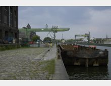 Industriehafen mit alten Lagergebäuden aus dunklem Backstein auf der linken Seite, einem Kopfsteinpflasterweg und grünen Hafenkränen mit der Aufschrift "BEHALA". Rechts liegt ein rostiges Frachtschiff am Ufer, und im Hintergrund sind moderne Gebäude und Industriebauten sichtbar.