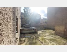 Hinterhof eines alten Gebäudekomplexes mit grober Putzfassade, bröckelnden Wänden und einem mit Moos bewachsenen Betonboden. Im Hintergrund stehen Bäume, eine niedrige Belüftungsanlage und weitere Gebäude mit spärlichen Fenstern.