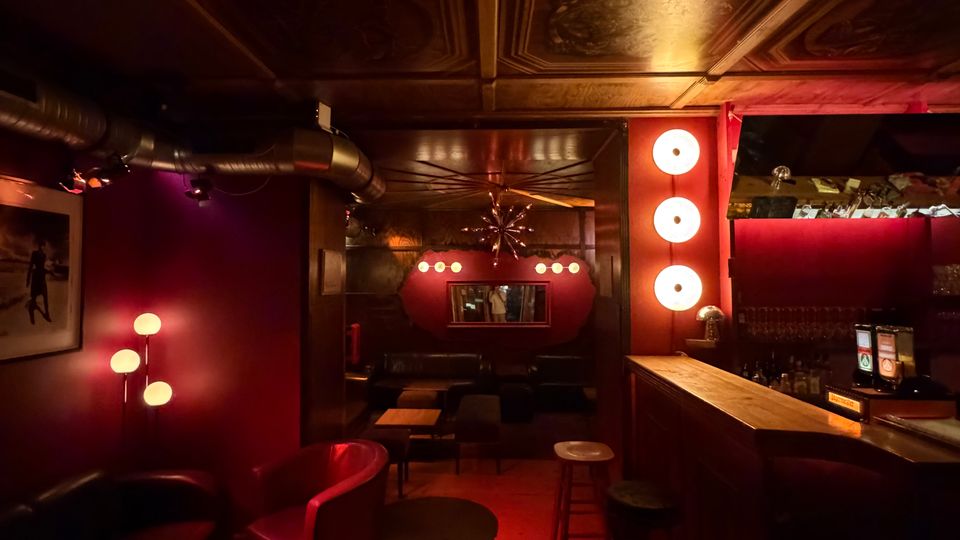 Innenraum einer stilvollen Bar/Lounge mit rot gedämpfter Beleuchtung, gepolsterten Sitzbänken und kleinen Tischen sowie einem langen Tresen mit Barhockern. Auffällig sind die dekorierte Kassettendecke, runde Wandlampen und ein spiegelnder Bereich im Hintergrund.