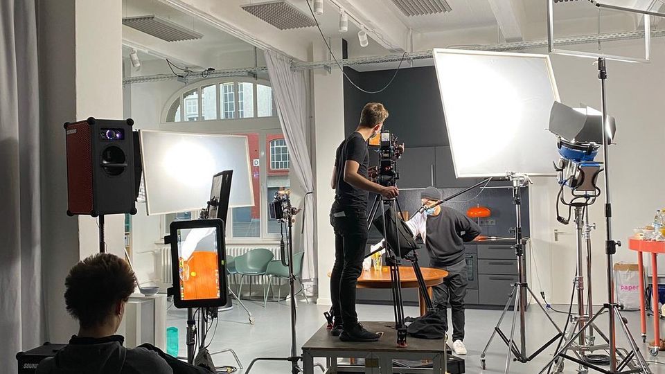Innenaufnahme eines hellen Fotostudios mit hohen Decken, sichtbaren Trägern und akustischen Paneelen, ausgestattet mit großen Softboxen, Scheinwerfern auf Stativen, Kameraaufbau und Kabeln. Im Hintergrund stehen ein Tisch vor einer dunklen Küchenzeile sowie große Fenster und Vorhänge.