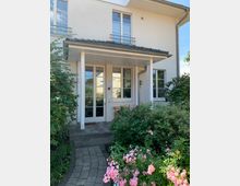 Eingang eines modernen Wohnhauses mit heller Fassade, überdachter Haustür und großen Fenstern. Vor dem Eingang führt ein gepflasterter Weg durch einen gepflegten Garten mit blühenden Sträuchern und rosa Blumen.