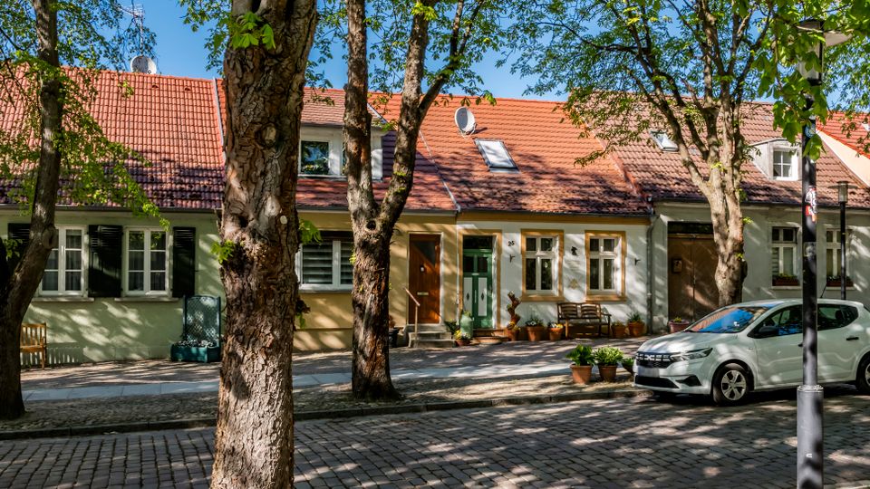 Tree-lined cobblestone residential street with a row of small pastel houses featuring red tiled roofs, shuttered windows, and front doors facing the sidewalk. Potted plants and benches sit along the facades, and a small white car is parked at the curb under leafy trees.
