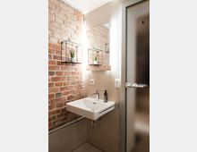 Kleines Badezimmer mit einer freistehenden weißen Waschschüssel und Metallarmaturen vor einer Wand aus sichtbaren Ziegelsteinen. Rechts befindet sich eine gerahmte Glastür mit mattierter Oberfläche, und neben dem Waschbecken ist ein beleuchteter Spiegel angebracht.