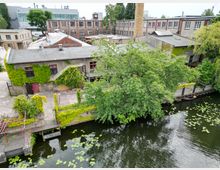 Industriegebäude an einem Fluss mit dichter Begrünung, darunter Efeu an den Fassaden. Im Vordergrund eine Uferpromenade mit Holzstegen und Wasserpflanzen auf der Wasseroberfläche, im Hintergrund größere Backstein- und Fabrikgebäude.