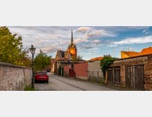 Pflastersteinstraße in einem Dorf mit Backsteinmauern und alten Scheunentoren auf der rechten Seite, Bäumen und einer Straßenlaterne links; im Hintergrund ist eine Kirche mit spitzem Turmdach zu sehen, beleuchtet vom warmen Licht der untergehenden Sonne.