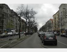 Städtische Straße mit mehrstöckigen Wohngebäuden auf beiden Seiten, kahlen Bäumen entlang des Gehwegs und geparkten Autos am Straßenrand; über dem Bild ein bewölkter Himmel.