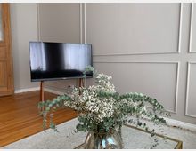 Wohnzimmer mit einem Flachbildfernseher auf einem Holzständer, hellem Holzboden, einer cremefarbenen Wand mit dekorativen Rahmenleisten und einem Couchtisch mit einem Blumenstrauß aus Eukalyptus und weißen Blüten in einer Vase.
