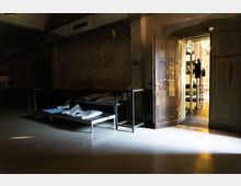 Dunkler, karger Innenraum mit rohen Betonwänden und sichtbaren Lüftungskanälen an der Decke, in dem zwei einfache Metallbetten mit Kissen stehen. Eine offene, abgenutzte Holztür führt in einen helleren Nebenraum mit industriellen Regalen und Geräten.