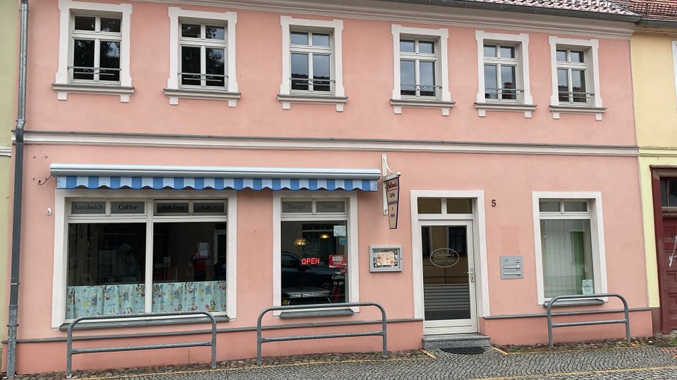 Zweistöckiges, rosafarbenes Stadthaus mit roten Ziegeln, weißen Fensterrahmen und mehreren Sprossenfenstern; im Erdgeschoss befindet sich ein kleines Café mit blau-weiß gestreifter Markise und großen Schaufenstern. Vor dem Gebäude verläuft ein gepflasterter Gehweg mit niedrigen Metallbügeln entlang der Straße.