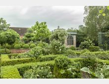 Ein gepflegter Garten mit dicht geschnittenen Hecken, Buchsbaumkugeln und blühenden Rosen. Im Hintergrund befindet sich ein kleines Backsteingebäude mit bogenförmigen Fenstern, umgeben von Bäumen und einer Gitterkonstruktion.