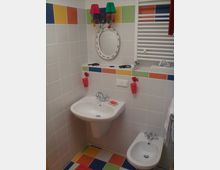 Bunt eingerichtetes Badezimmer mit weiß gefliesten Wänden und farbigen Kacheldetails in Rot, Grün, Gelb und Blau. Zu sehen sind ein Waschbecken mit Spiegel und farbiger Wandlampe darüber, ein Bidet sowie ein Handtuchhalter und ein Heizkörper an der Wand. Der Boden ist mit Kacheln in denselben Farben gefliest.