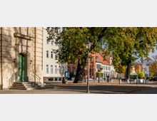 Eine städtische Straße mit historischen Gebäuden, darunter ein sandfarbenes Haus mit grünem Eingang und Stufen im Vordergrund. Auf der rechten Seite stehen Bäume mit gelb-grünem Herbstlaub, Straßenlaternen und Bänke, die den Platz beleben.