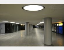 Unterirdischer Durchgang mit Betonpfeilern, großen runden Deckenleuchten und einem Boden aus grauem Beton. An den Wänden sind Türen, Schaufenster und Werbetafeln zu sehen, im Hintergrund stehen zwei Personen.