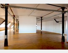 Großer, offener Innenraum mit hellen Wänden, einem Boden aus Holzparkett und mehreren schwarzen Stahlträgern, die die Decke stützen. Die Decke ist flach und mit Lampen und Kabeltrassen ausgestattet, während Fenster auf der linken Seite Tageslicht einlassen.