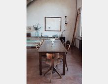 Ein minimalistischer Essbereich mit einem langen Holztisch und fünf unterschiedlichen Holzstühlen. Im Hintergrund eine helle Wand mit schlichten Dekoelementen, darunter ein Regal mit Büchern und Geschirr, eine Leiter und eine Wandleuchte.