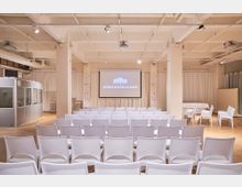 Veranstaltungsraum mit weißem Holzboden, bestuhlt mit weißen Stühlen in Reihen vor einer Leinwand mit der Aufschrift „SPREESPEICHER“. Im Hintergrund sind helle Vorhänge, eine Dolmetscherkabine und ein Projektor an der Decke sichtbar.