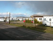 Ein Campingplatz mit mehreren geparkten Wohnwagen, umgeben von einem einfachen Maschendrahtzaun. Im Hintergrund befindet sich ein flaches Gebäude mit einem Satteldach, und links ist ein kleiner Eingang mit einem Schild sichtbar.