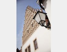 Fassadendetail eines historischen Gebäudes mit heller, verputzter Oberfläche und auffälligem Treppengiebel aus geometrischen Bögen; im Vordergrund eine schmiedeeiserne Laterne.