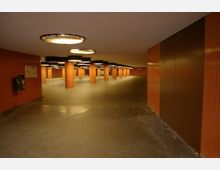 Unterirdischer Durchgangsbereich mit orangefarbenen Fliesenwänden, mehreren quadratischen Säulen und leuchtenden Deckenlampen. Der Boden ist mit dunklen Fliesen bedeckt, und an einer Wand hängt ein Abfallbehälter.